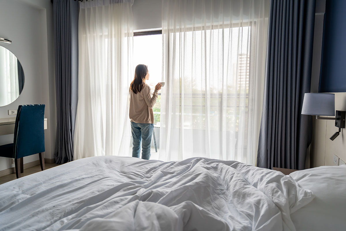 Young Asian Woman Holding Cup Of Coffee And Opening Curtains In The Morning And Looking Through The Window At Home